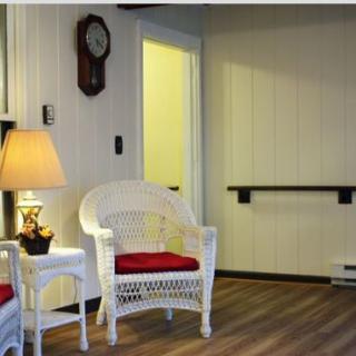 A cozy room featuring a wicker chair with a red cushion, a side table with a lamp, and a wall-mounted clock.
