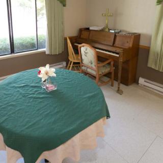 A room with a piano and chair in the corner, a round table with a green tablecloth, and a flower arrangement on top.