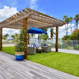 A sunny outdoor seating area with a pergola and umbrella on a wooden deck, surrounded by a garden and palm trees.