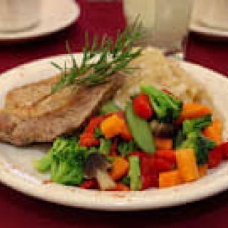 A plated meal featuring a pork chop with a side of mixed vegetables and mashed potatoes, garnished with rosemary.