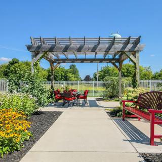 A scenic garden with a pergola and outdoor seating area surrounded by blooming flowers on a sunny day.