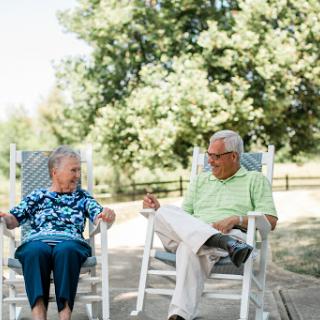 An elderly couple enjoying a sunny day together on rocking chairs, sharing smiles and conversation in a serene outdoor setting.