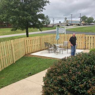 A person standing on a small patio enclosed by a wooden fence, with chairs and outdoor scenery.