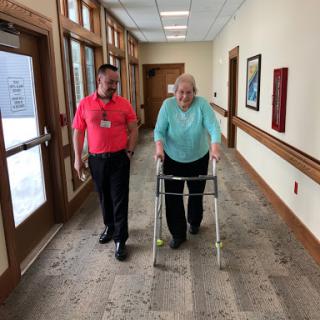 A caregiver assists an elderly woman using a walker in a bright corridor.