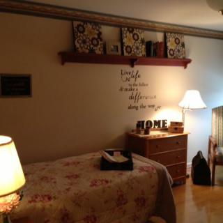 A cozy bedroom with warm lighting, featuring a bed with floral bedding, decorative wall art, and a wooden dresser.