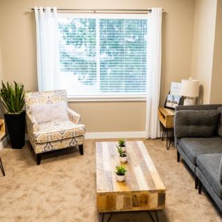 A cozy living room with a gray sofa, patterned armchair, wooden coffee table, and a plant near the window.