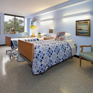 A hospital room featuring a neatly made bed with a patterned quilt, a window with balloons, and comfortable seating.