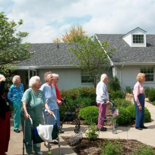 A group of elderly individuals walking outdoors in a garden setting at a retirement home.