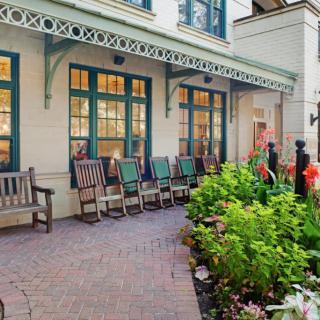A cozy outdoor patio area with benches and rocking chairs, surrounded by lush greenery and a charming building facade.
