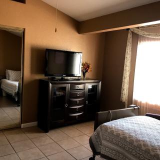 A cozy bedroom featuring a TV on a dresser, with a mirror and window adorned with curtains.