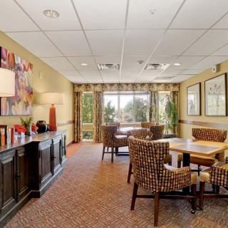 A cozy dining area with a table and chairs, decorated with artwork and lamps, offering a view through large windows.
