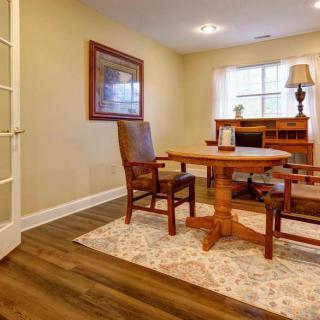 A cozy home office with a wooden desk and chairs, featuring a rug and a lamp by the window.
