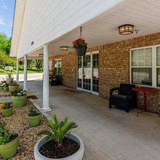 A covered porch with brick walls, featuring potted plants, patio furniture, and decorative columns.