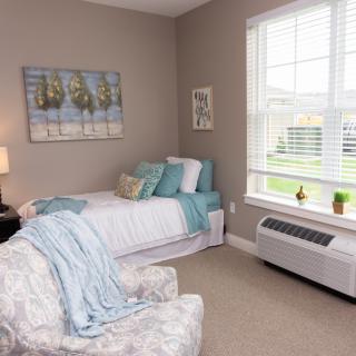A cozy, modern bedroom with a single bed adorned with blue and white linens, complemented by a comfortable armchair and wall art, bathed in natural light from a large window.