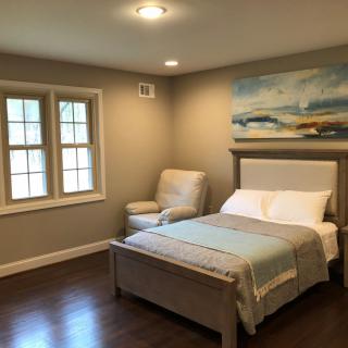 A cozy bedroom featuring a neatly made bed, an armchair by the window, and a piece of wall art above the headboard.