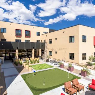 A modern building with a courtyard featuring a small lawn and seating area under a blue sky.