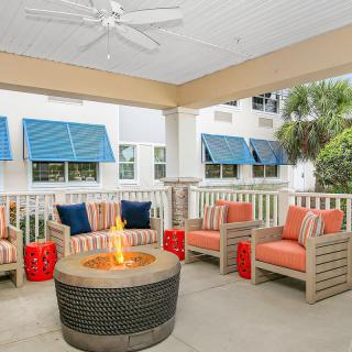 A cozy outdoor patio area with cushioned chairs around a fire pit, shaded by a ceiling fan and decorated with colorful awnings.