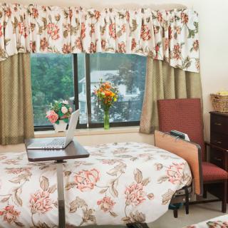 A cozy bedroom with floral decor, featuring a laptop on the bed, and a scenic window view.