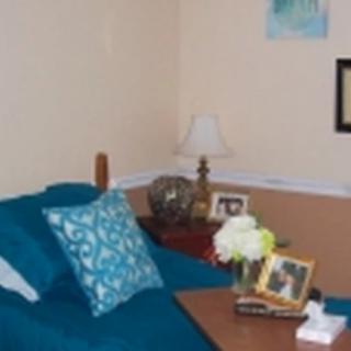 A cozy living room setup with a teal couch, decorative pillow, and a table adorned with flowers, a lamp, and framed photos.