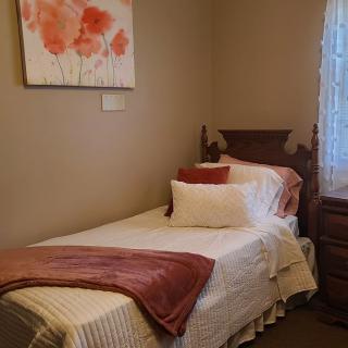 A cozy bedroom with a single bed, adorned with pillows and a blanket, featuring artwork on the wall and natural light from a window.