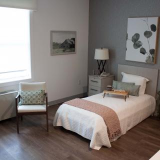 A modern, minimalist bedroom featuring a single bed with a simple yet elegant design, complemented by a chair and artwork on the walls.