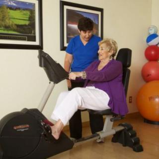 A woman assists an older adult with physical therapy exercises using specialized fitness equipment.