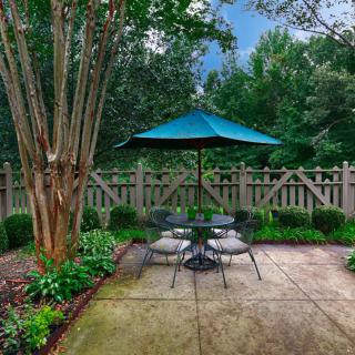A cozy backyard patio with a table, chairs, and an umbrella surrounded by lush greenery.
