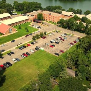 An aerial view of a facility with a parking lot, surrounded by greenery and a river.