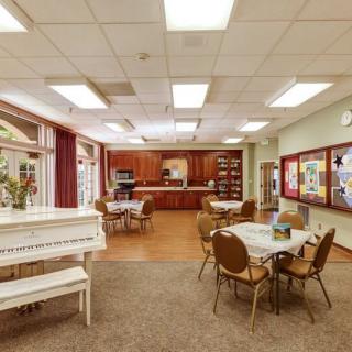 A cozy communal room featuring a white piano, tables with chairs, and a kitchenette area.
