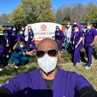A group of healthcare workers in purple uniforms take a selfie outdoors, all wearing masks.