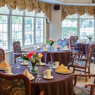 A cozy dining area with elegantly set tables and chairs, adorned with floral centerpieces near large windows.