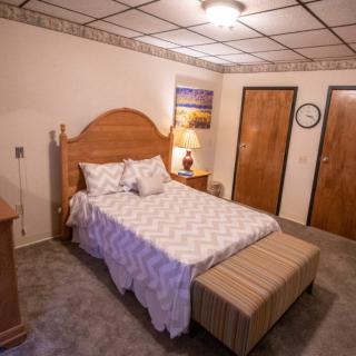 A cozy bedroom with a wooden bed frame, patterned bedding, and a striped bench at the foot of the bed. The room includes a lamp on a nightstand and two wooden doors.