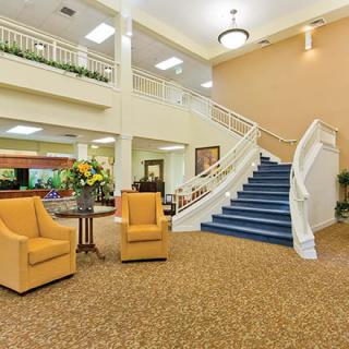 A spacious lobby featuring a carpeted floor, comfortable seating arrangements, a prominent staircase, and decorative flowers.