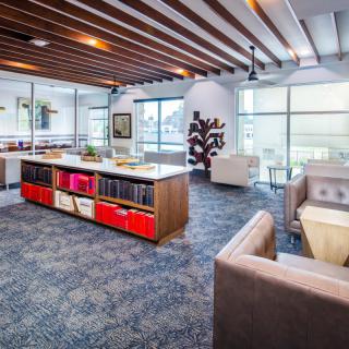 A modern office space with open layout, featuring wooden ceiling beams, stylish furniture, bookshelves, and ample natural light.