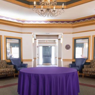 An elegantly decorated room featuring a purple tablecloth-covered table, surrounded by seating and lit by a chandelier.