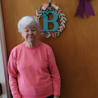 An elderly woman stands by a decorative door, showcasing a warm smile in a cozy indoor setting.