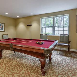 A cozy billiards room featuring a pool table with red felt and classic wooden chairs, illuminated by natural light from a large window.