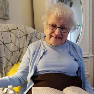 An elderly woman smiles while sitting comfortably in a chair with an open book.