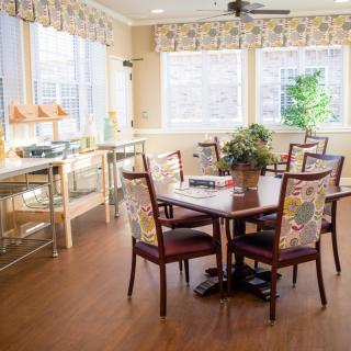 A well-lit dining room with a round table surrounded by chairs, featuring large windows and a hardwood floor.