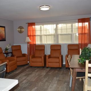 A cozy living room with several brown recliner chairs, a large window with orange curtains, and a wooden table decorated with plants.
