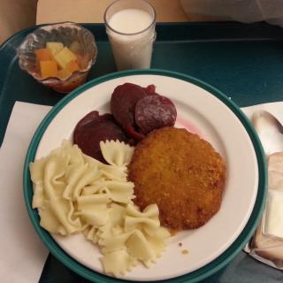 A meal served on a tray includes pasta, a fried cutlet, sliced beets, fruit salad, and a glass of milk.
