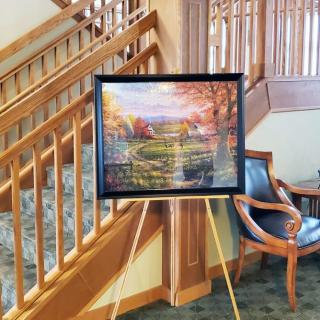 A framed painting displayed on an easel in a warmly lit interior with a wooden staircase and chair in the background.
