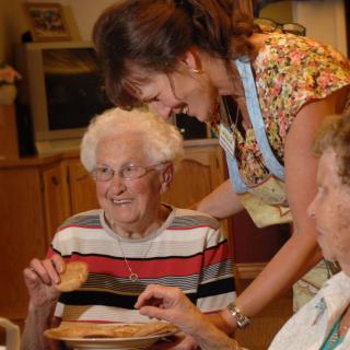 A caregiver assists elderly individuals during a meal in a communal setting.