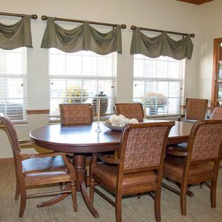 A cozy dining room with a wooden table and six chairs, framed by three large windows with green curtains.