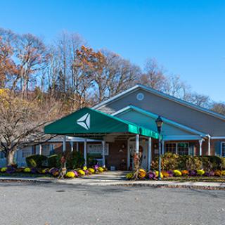 A building with a green awning is surrounded by autumn trees and plants, featuring a parking lot in the foreground.
