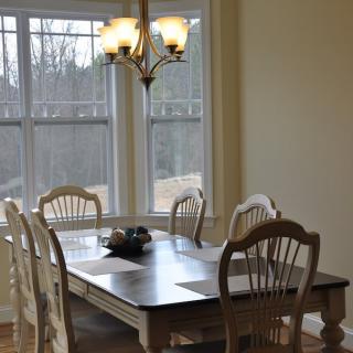 A cozy dining area with a wooden table and chairs, illuminated by a chandelier next to large windows.