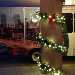 A festive display featuring Christmas garland and lights wrapped around a pillar, with a decorated bus in the background.