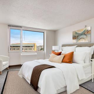 A modern bedroom with neutral tones, featuring a double bed adorned with orange and brown pillows, a large window with a city view, and elegant decor.