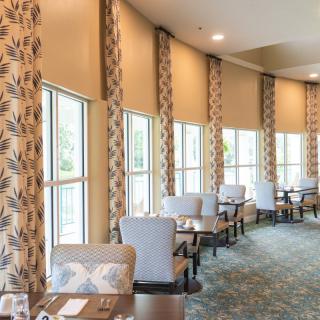 A well-lit dining area featuring tables and chairs, with patterned curtains and large windows.