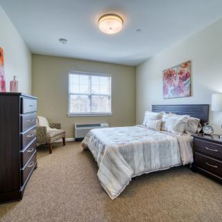 A cozy bedroom with a bed, dresser, and chair, featuring warm lighting and neutral decor.